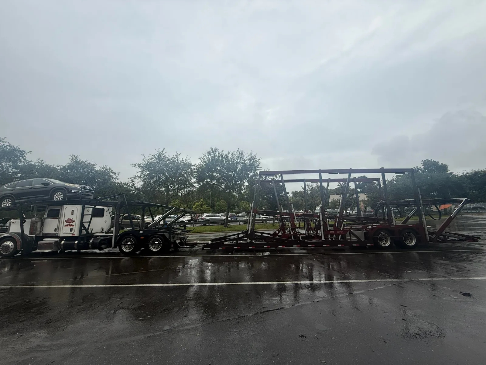 Open car carrier with an SUV on the upper rack, loading at a vehicle lot on a rainy day