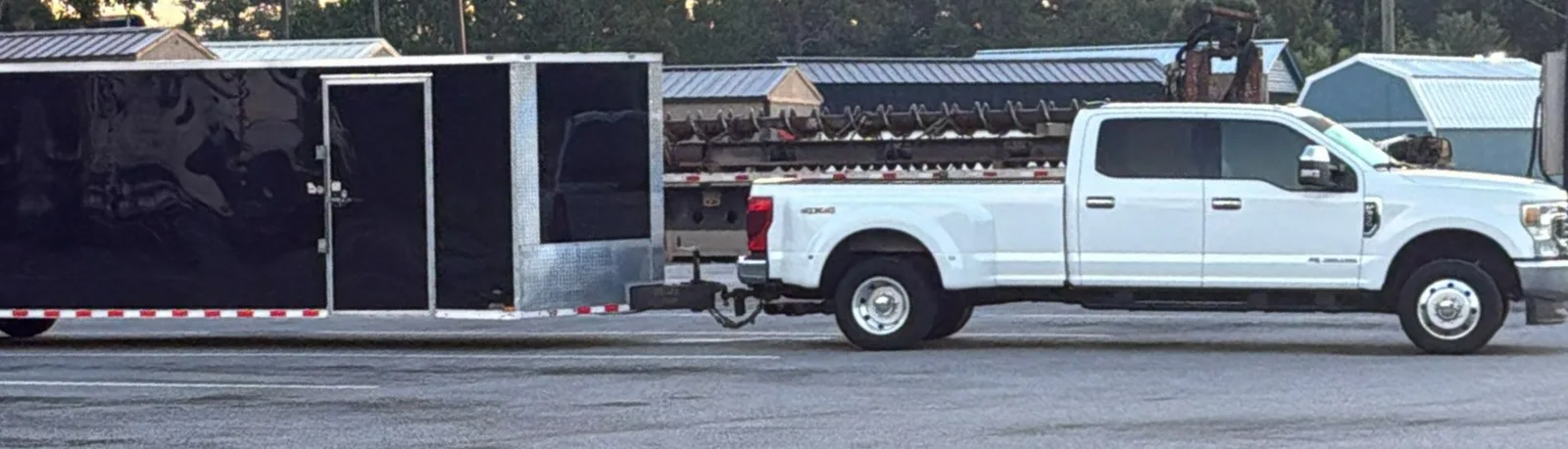 White dually pickup towing a black enclosed car trailer across a parking lot at sunset