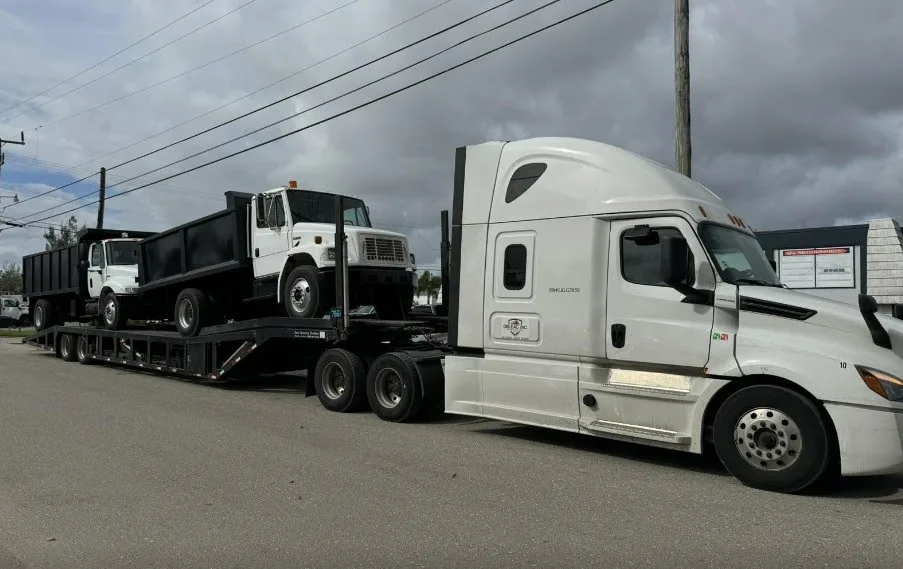 Two dump trucks loaded on a step-deck trailer behind a white semi-truck for heavy equipment transport